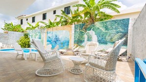 Un área junto a la piscina con sillas de mimbre blanco y una mesa, rodeada por un mural con motivos tropicales.