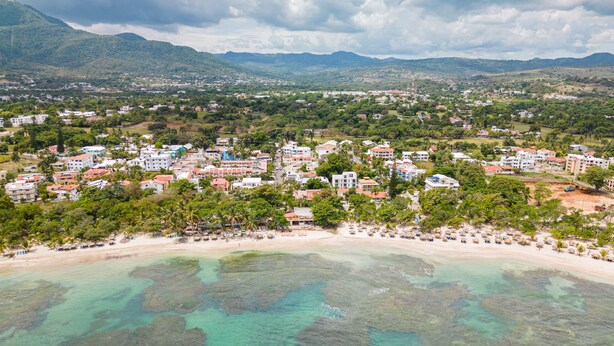 Vista aérea de un pueblo costero con edificios, una playa y montañas al fondo.