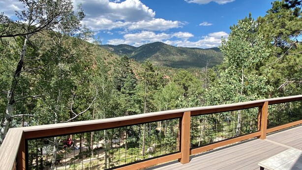 Una terraza de madera con baranda de vidrio que da a un valle boscoso y montañas.