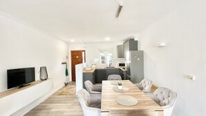 Ein modernes Wohnzimmer mit einem Holzesstisch, weißen Stühlen, einem Flachbildfernseher und einer Küche im Hintergrund.