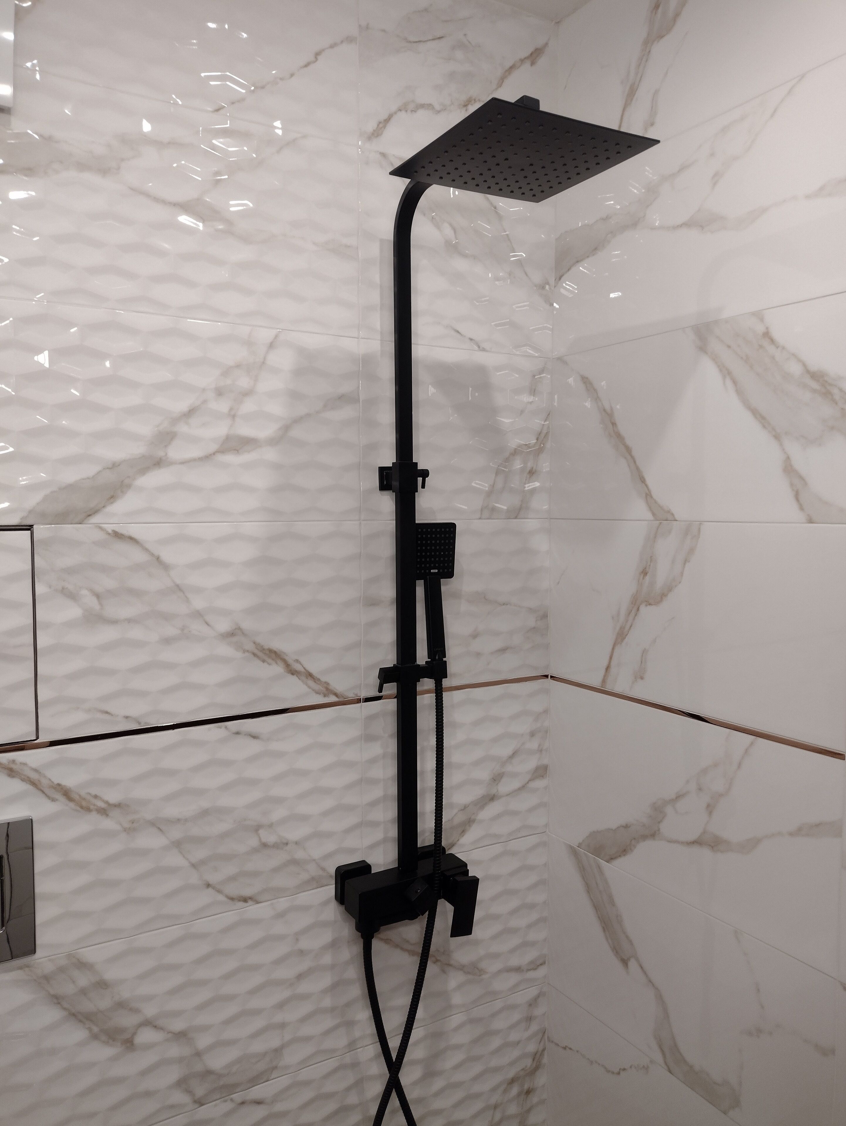 A shower with a handheld showerhead mounted on a black stand, set against a white marble wall with a textured pattern.