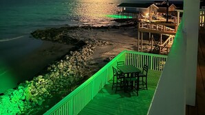 A nighttime view of a beachside resort with a wooden deck, a small table and chairs, and a distant view of the ocean under a bright moon. - Beachfront Luxury Retreat – Oceanfront, Family & Pet-Friendly Paradise (Freeport)