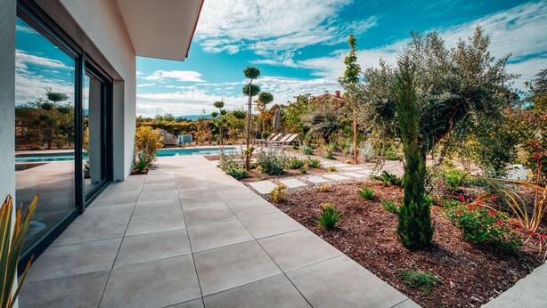 A modern house with a large glass window, a patio with a pool, and a garden with various plants.