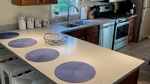 A kitchen with wooden cabinets, a stainless steel refrigerator, a microwave, and a stove. There are four bar stools with woven seats and a central island with a sink and a vase of flowers.