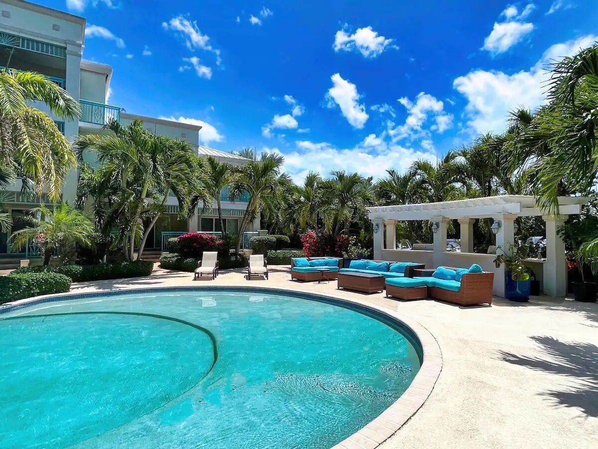 A pool area with lounge chairs and a covered seating area, surrounded by palm trees and a clear blue sky.