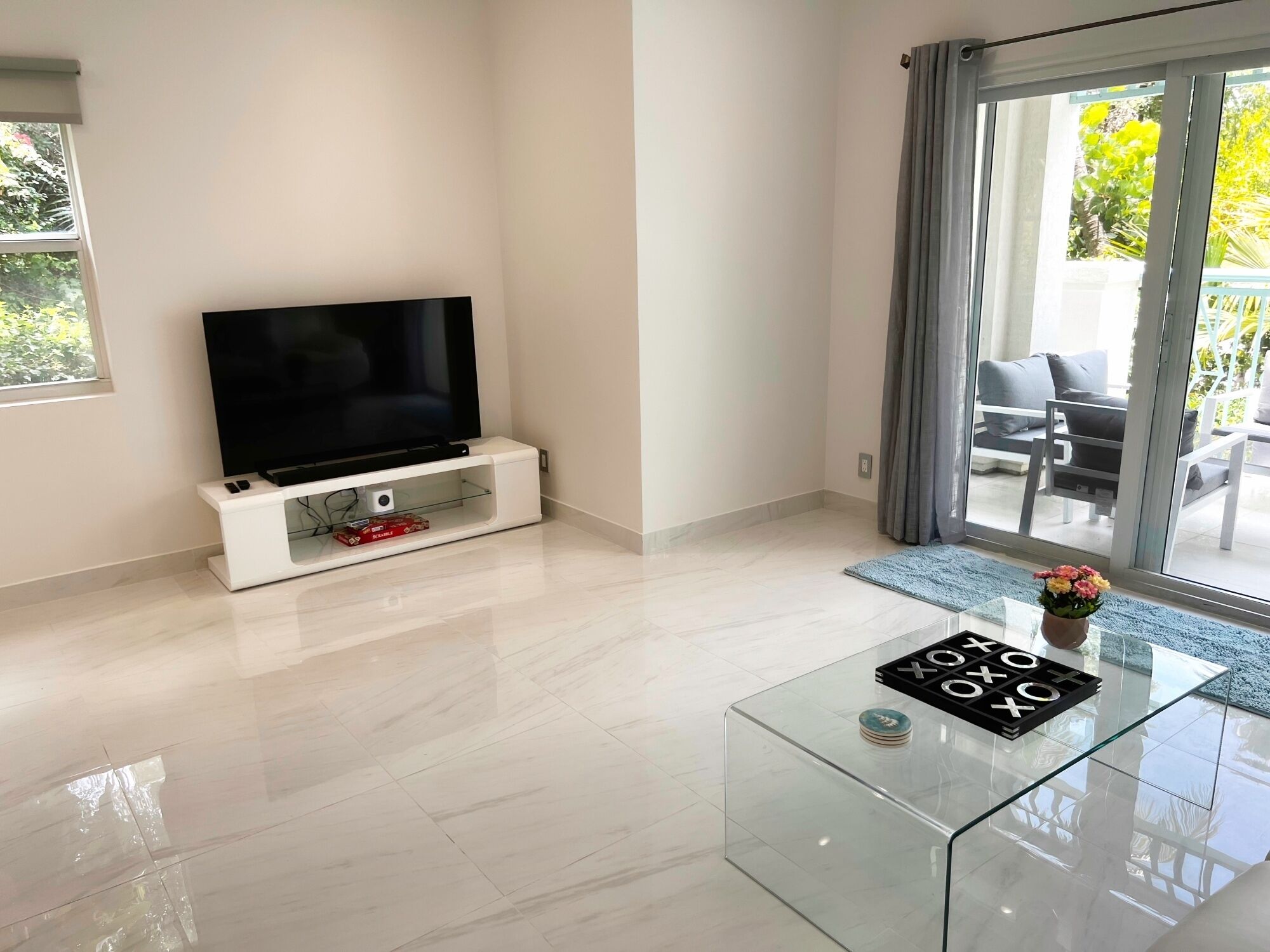 Un soggiorno moderno con una televisione a schermo piatto su un mobile bianco, un tavolino da caffè in vetro con un gioco da tavolo, e vista su un balcone con mobili da esterno.