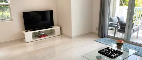 Un soggiorno moderno con una televisione a schermo piatto su un mobile bianco, un tavolino da caffè in vetro con un gioco da tavolo, e vista su un balcone con mobili da esterno.