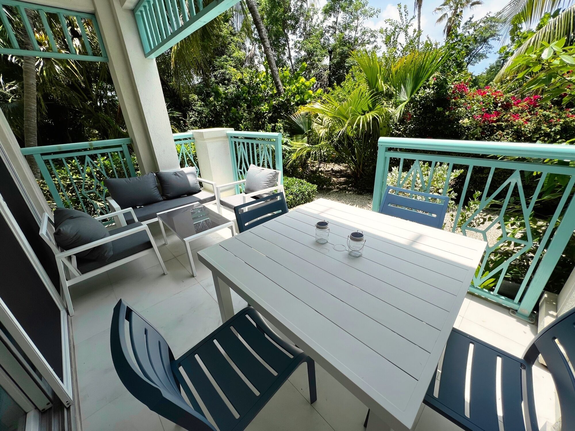 An outdoor patio with a white table and chairs, surrounded by lush greenery and a blue railing.