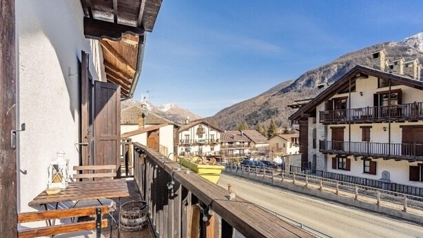 A balcony with wooden furniture, a view of a mountain, and traditional buildings in the background. - The Loving Cabin  (Pragelato-Ruà)