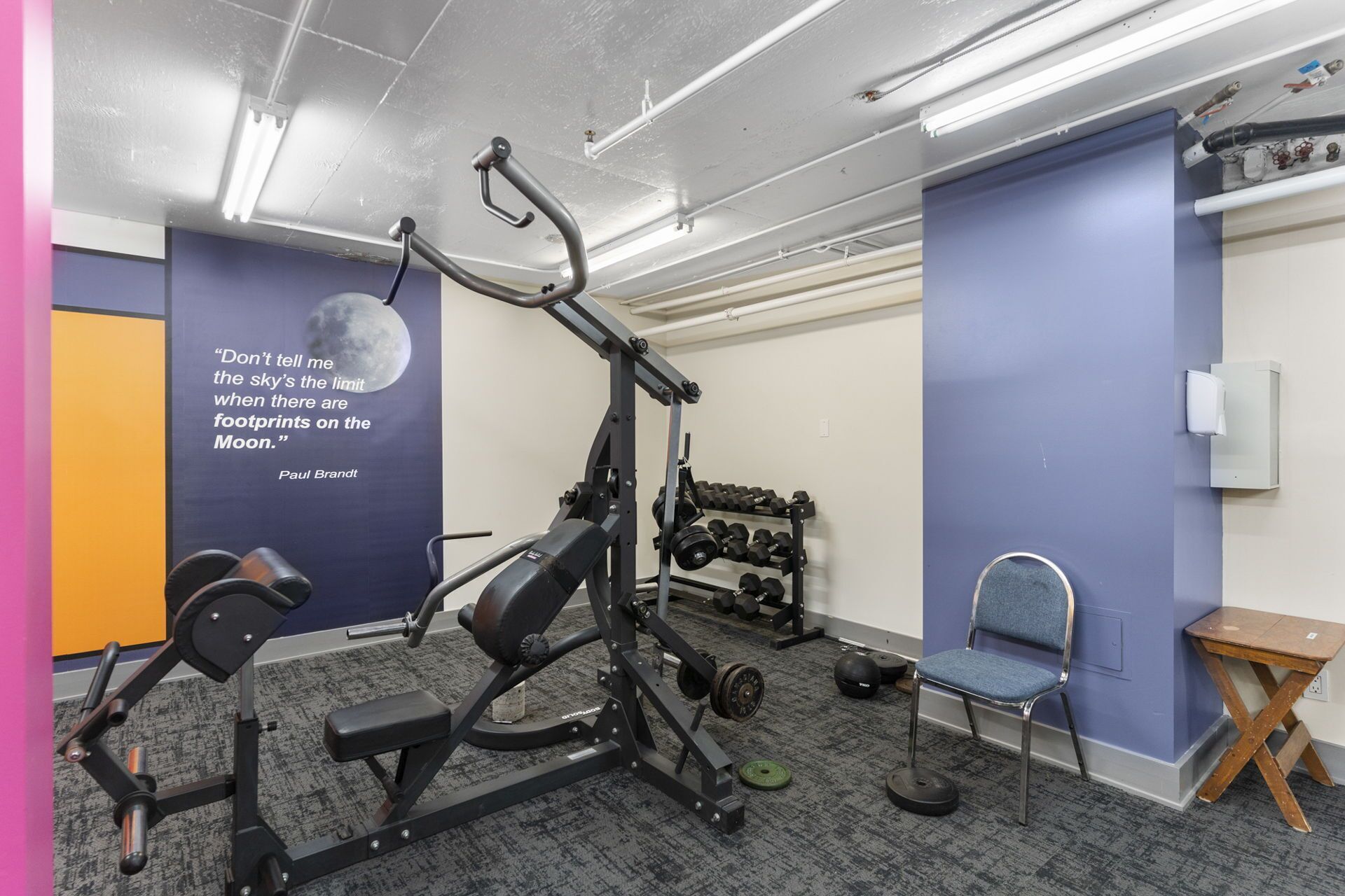 Een sportschool met trainingsapparatuur, een motiverende quote op de muur en een stoel.