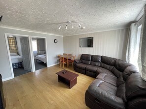 A living room with a brown leather sofa, a wooden coffee table, a dining table with chairs, a TV mounted on the wall, and a bed in the adjacent room.