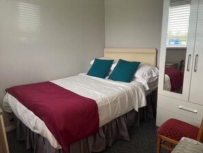 Una cama individual con ropa de cama blanca y turquesa, una manta roja y un mueble espejo con un cojín rojo.