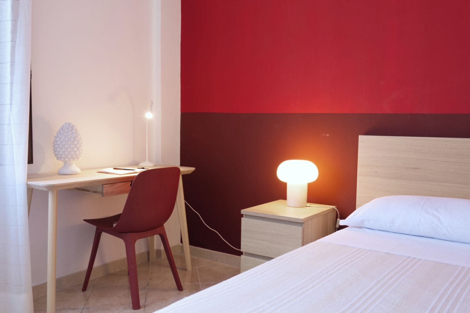 A hotel room with a desk, chair, bedside table with a lamp, and a bed.