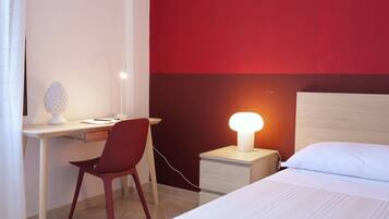 A hotel room with a desk, chair, bedside table with a lamp, and a bed.