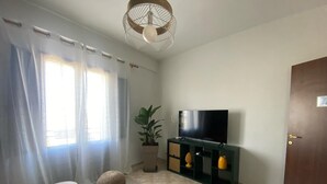 A living room with a flat-screen TV on a stand, a potted plant, a round ottoman, and a door. - Le Stanze di Archimede (Syracuse)
