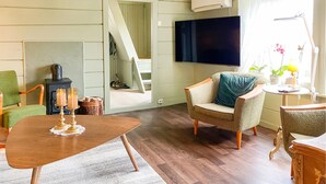 Un salon chaleureux avec une table en bois, un fauteuil vert, un fauteuil beige avec un coussin bleu, un téléviseur et une petite table sur laquelle se trouvent une lampe et des plantes.