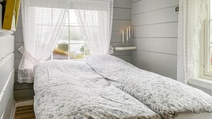 Une chambre à coucher avec un lit, une fenêtre ornée de rideaux en dentelle, une table de chevet avec des bougies et une vue sur l’extérieur.