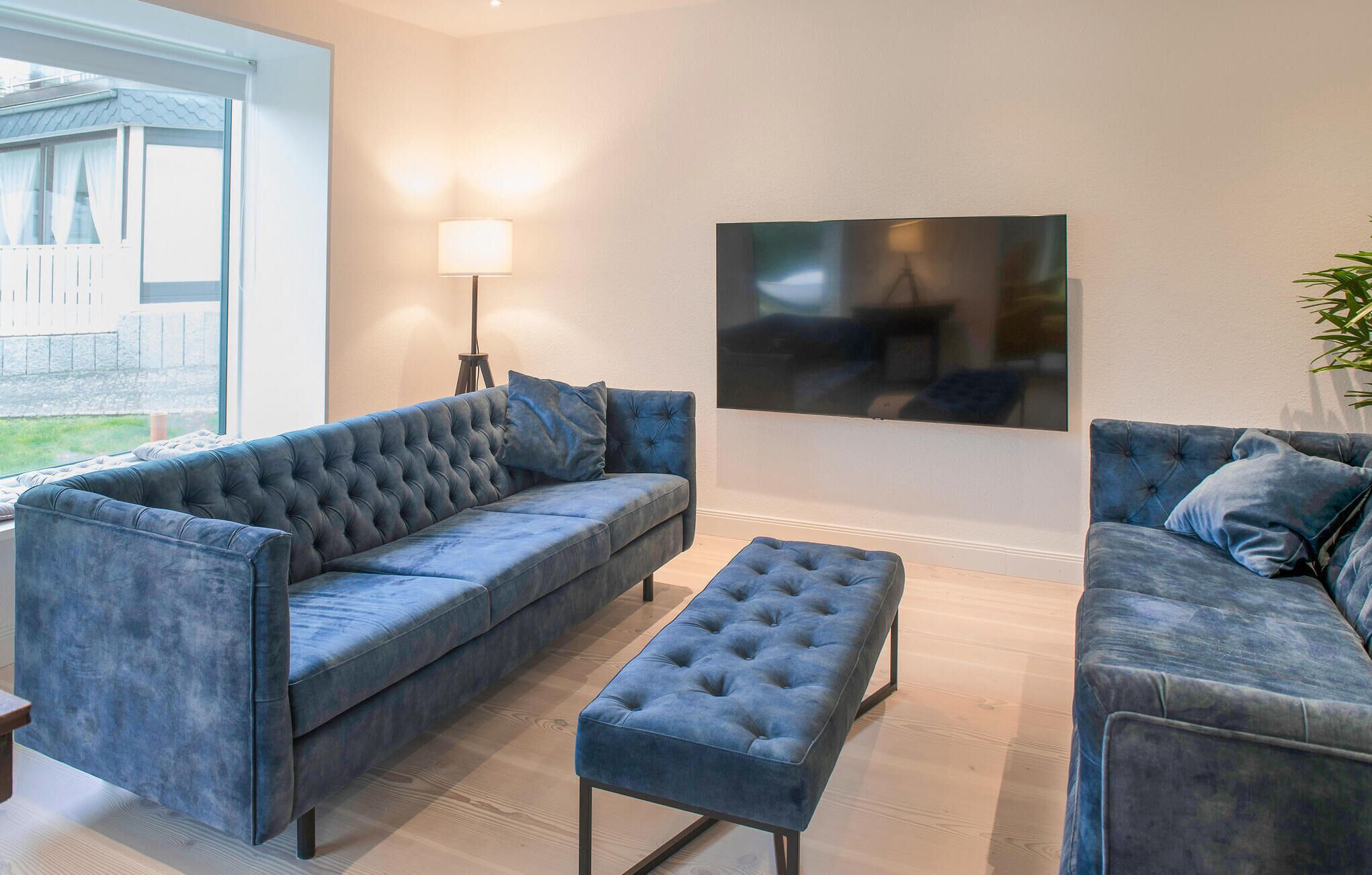 Een woonkamer met een grote bank, een kleinere bank en een poef, allemaal bekleed met dezelfde blauwe stof. Een staande lamp en een flatscreen-televisie zijn aan de muur bevestigd.