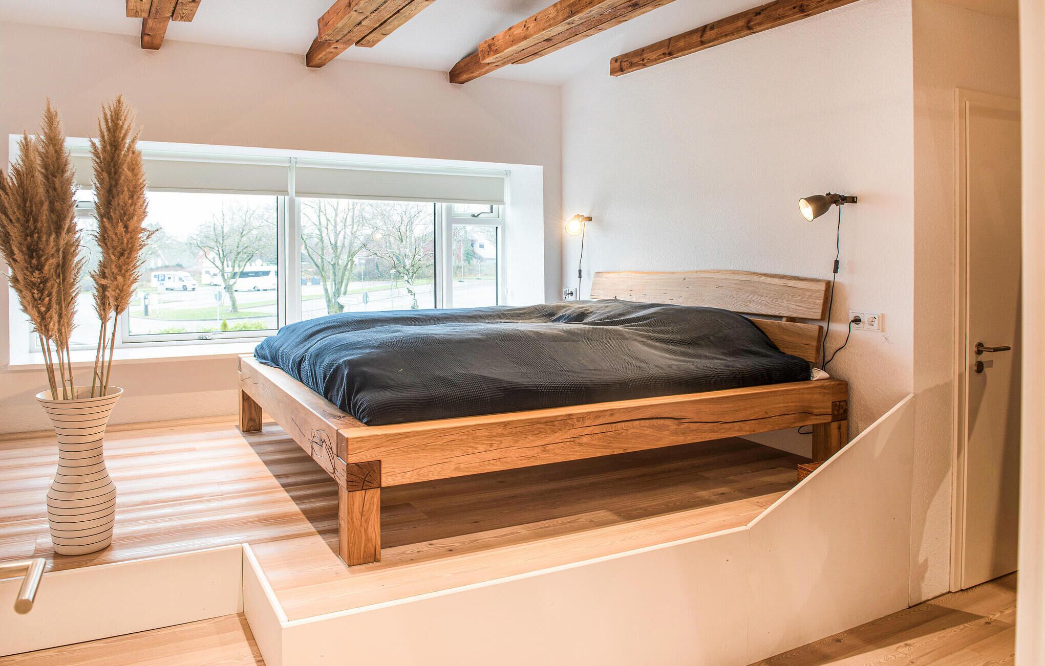 A bedroom with a wooden bed, a window with a view of the outside, and a vase with pampas grass.