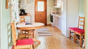 Una cucina compatta con mobili in legno, un fornello e un lavandino. C'è un tavolo da pranzo con due sedie, una con cuscino rosso. Una porta in legno dà accesso a un'altra stanza.