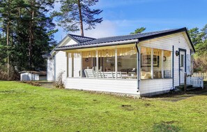 Ein kleines, eingeschossiges weißes Gebäude mit überdachter Terrasse, großen Fenstern und einem eingezäunten Garten.