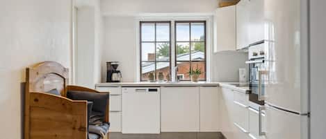 Eine kompakte Küche mit weißen Schränken, einem integrierten Geschirrspüler und einem Kühlschrank. Ein Fenster bietet Blick auf ein Backsteingebäude.