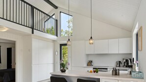 Une cuisine moderne avec des meubles blancs, un îlot central doté de deux chaises noires, des luminaires suspendus et une vue sur l’extérieur.