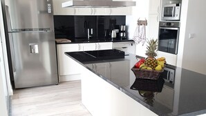 Una cuina moderna amb armaris blancs, electrodomèstics d'acer inoxidable i una illa central amb una cistella de fruita.