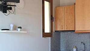 A compact kitchen with a refrigerator, wooden cabinets, a small dining table with chairs, and a TV mounted on the wall. - Kallia Apartment (Chania)