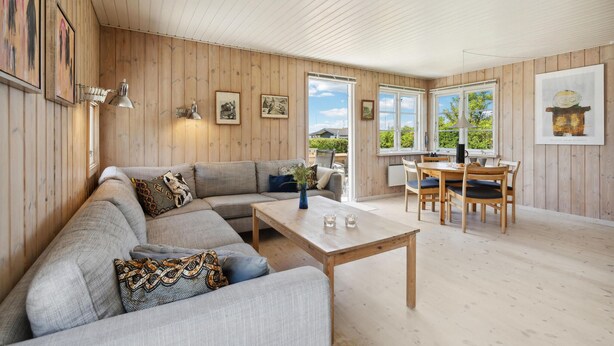 Amplia sala de estar con detalles de madera, un conjunto de sofás, una mesa de comedor con sillas y ventanas grandes que dan a vistas de vegetación.