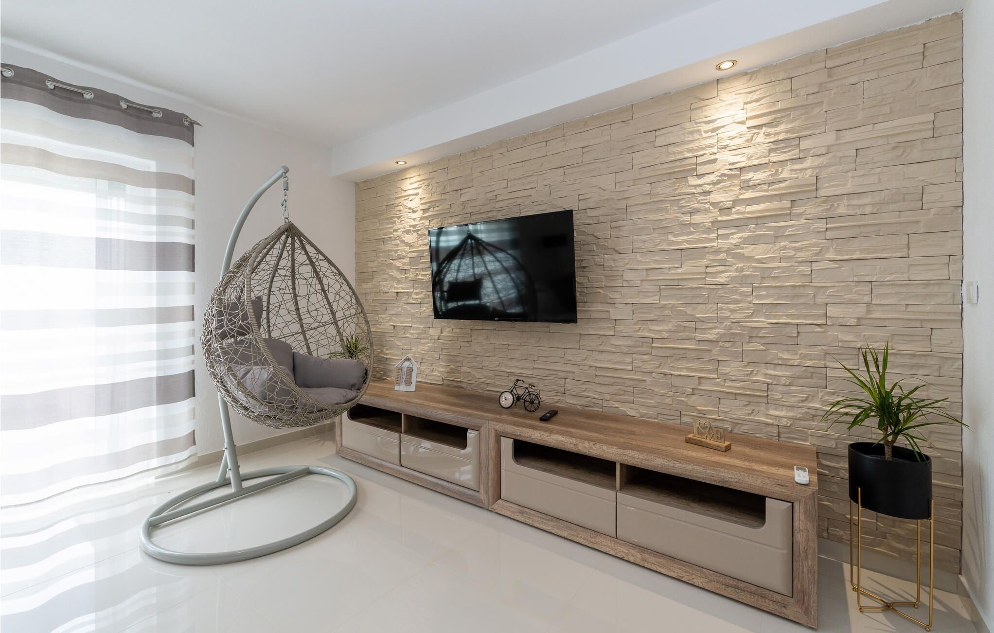A modern living room with a hanging chair, a flat-screen TV mounted on a textured wall, a wooden console table, and a potted plant.