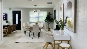 Uma sala de jantar moderna com uma mesa branca, cadeiras e um tapete. Há uma planta, um espelho fixado na parede e um quadro.