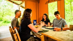 A group of people dining at a wooden table with a view of greenery outside. - Luxury Woodland Safari Tent with Private Hot Tub (Acton Scott)