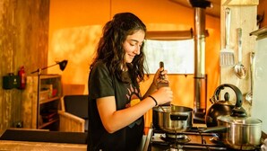 A person cooking in a kitchen with wooden walls, a gas stove, and various cooking utensils. - Luxury Woodland Safari Tent with Private Hot Tub (Acton Scott)
