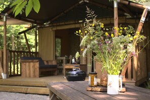 A rustic wooden table with a floral arrangement, jars, and a rustic pot in a covered outdoor area with wooden furniture and a wooden structure in the background. - Luxury Woodland Safari Tent with Private Hot Tub (Acton Scott)