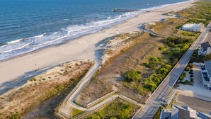 Vista aérea de una playa con un muelle de madera que se adentra en el océano, una playa de arena y una zona residencial con casas y una carretera pavimentada.
