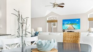 Una sala de estar moderna con comedor, que incluye un ventilador de techo, un televisor grande que muestra una escena tropical y un sofá blanco.