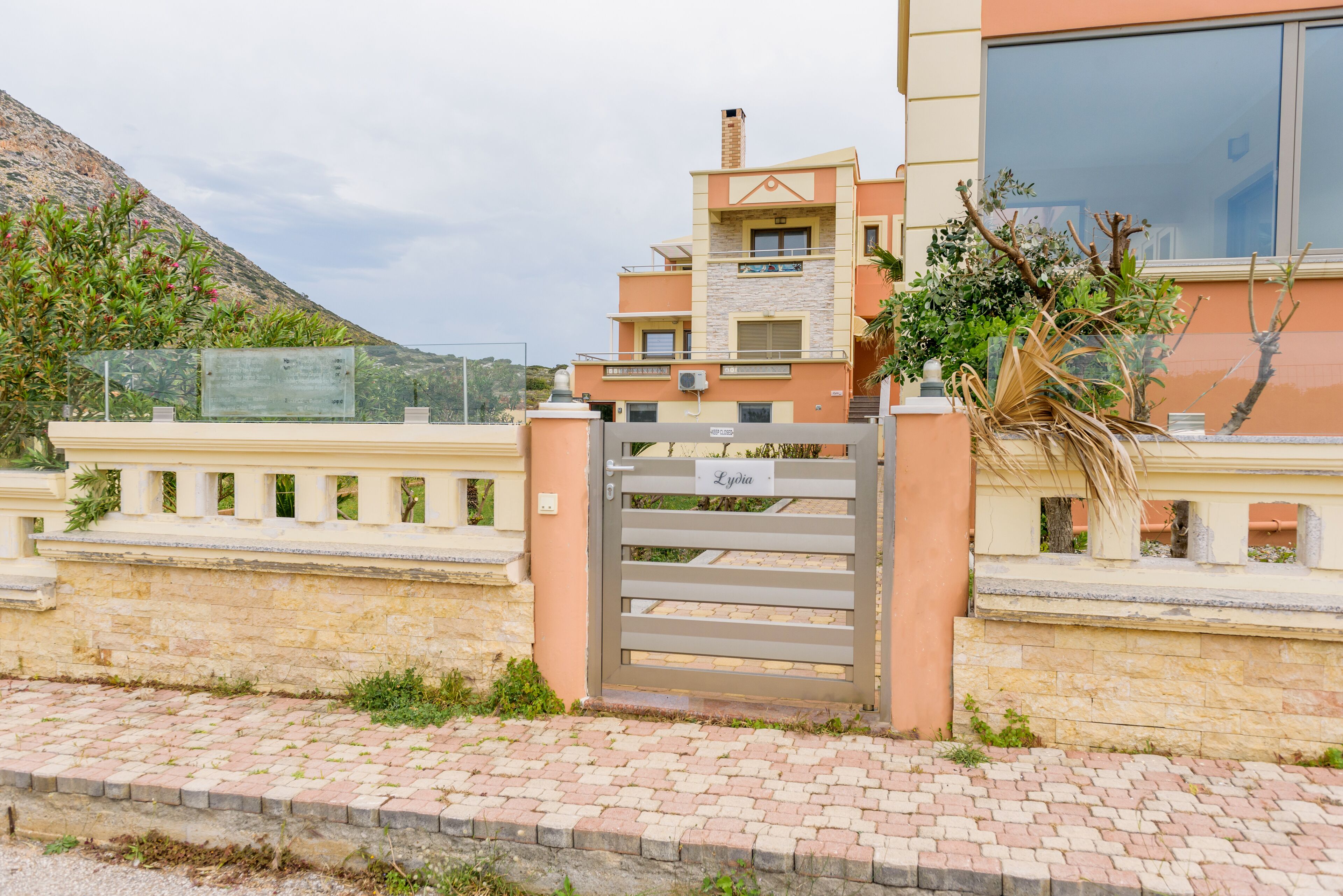A gated residential building with a stone wall, a paved entrance, and a mountain in the background.