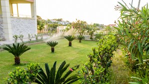 A patio with a white plastic chair and a glass table, surrounded by various plants and a building in the background. - Lydia Apartment (Chania)