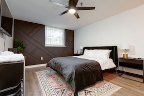 Un dormitorio moderno con una cama, ventilador de techo, ventana con persianas, mesita de noche con lámpara y una alfombra decorativa.