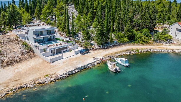 A modern multi-story house with a swimming pool, a boat docked at the water's edge, and a forested backdrop.