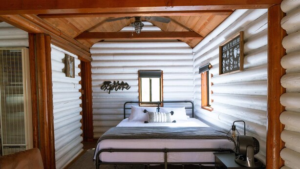 A log cabin room with a bed, a desk, a fan, and a window. - Hiker's Haven Summary (Mount Laguna)