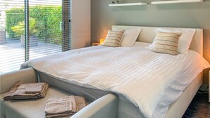 Un dormitorio moderno con una cama grande, dos almohadas, una mesita de noche con una lámpara y vistas a zonas verdes por la ventana.