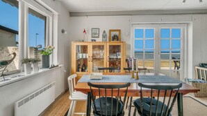 Uma sala de jantar com mesa e cadeiras de madeira, um armário com portas de vidro e vista para a praia através de grandes janelas.