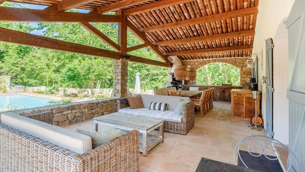 An outdoor patio with a stone wall, wicker furniture, a pool, and a stone archway leading to a kitchen area. - Awesome home in Carcès (Carcès)