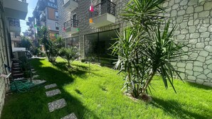 A courtyard with a stone wall, green grass, and small trees. There are steps leading to a door, and a hose is visible on the left. - Forbes Butik Otel (Izmir)