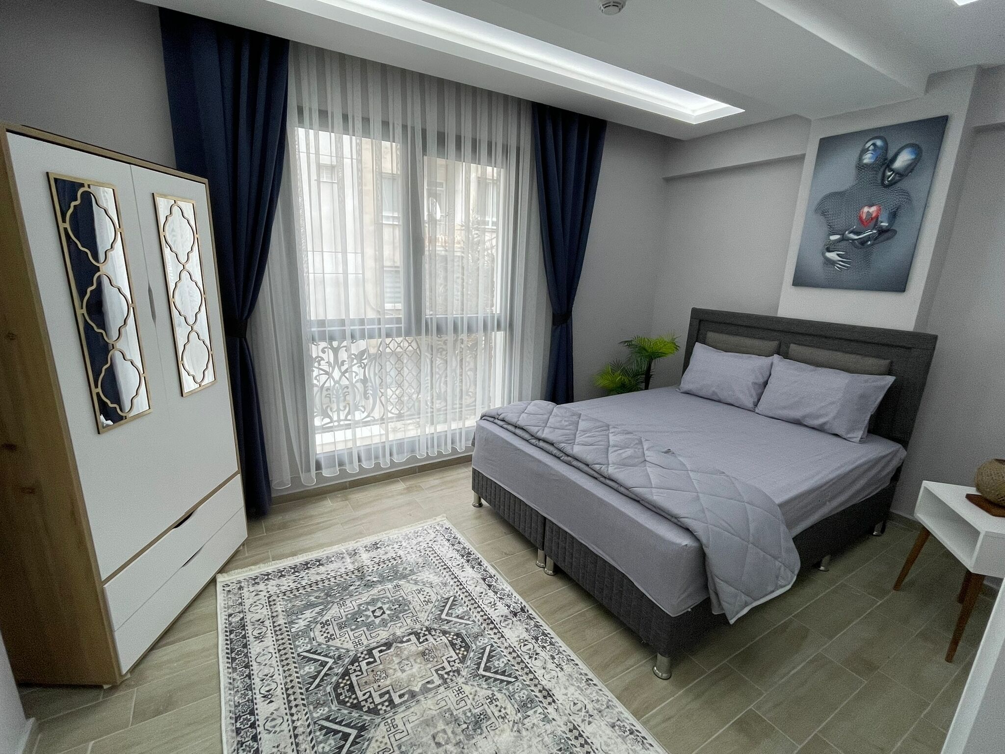 A modern bedroom with a bed, a wardrobe, a large window with curtains, and a painting on the wall.
