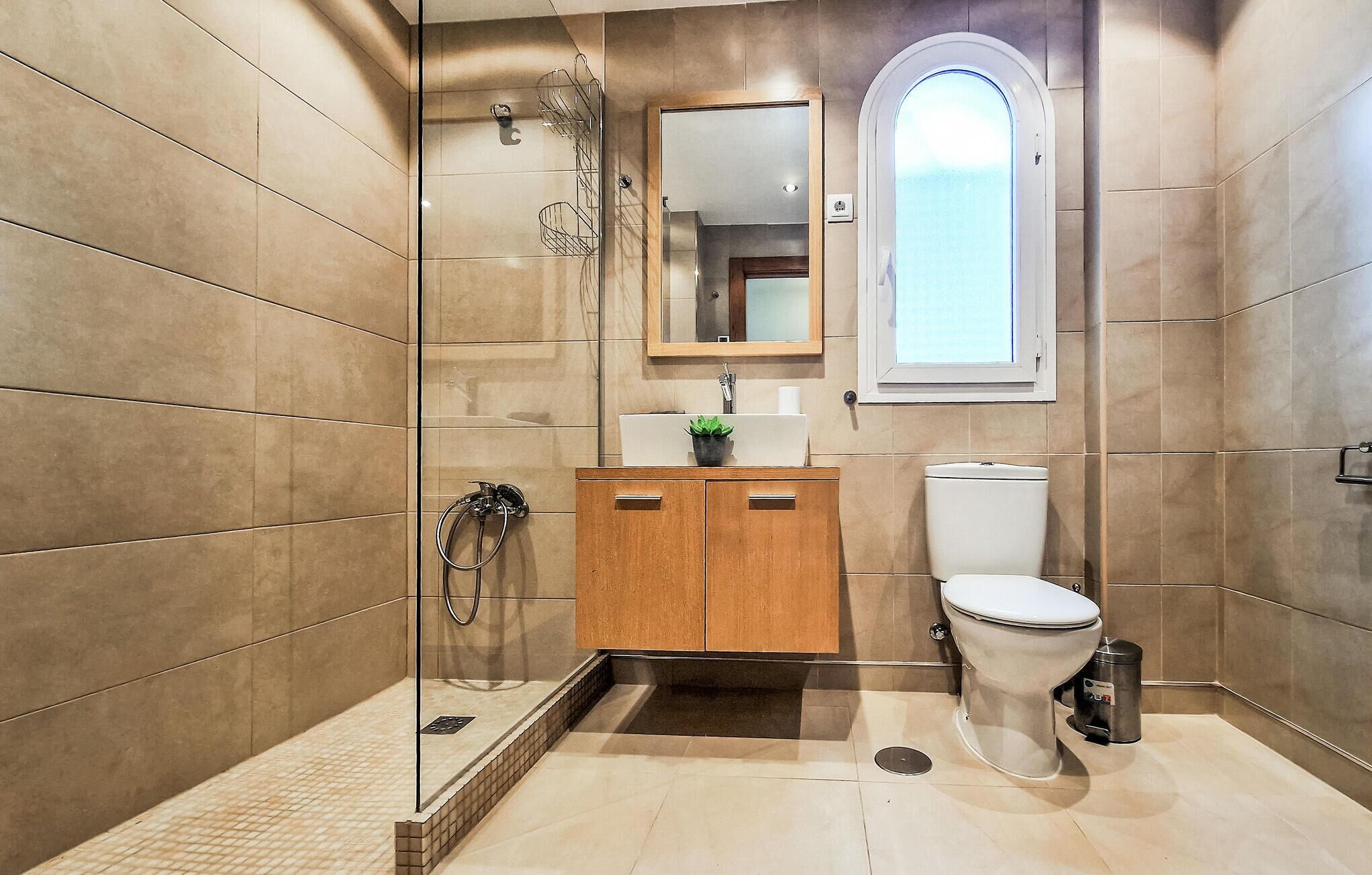 Un baño moderno con cabina de ducha de vidrio, tocador de madera, inodoro y una ventana pequeña.