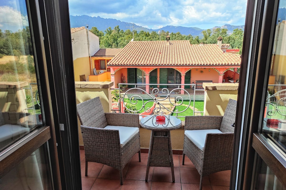 A balcony with wicker furniture and a table, overlooking a house with a tiled roof and a garden.