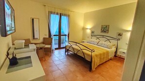A hotel room with a bed, a small table, a chair, a sofa, a window with curtains, and a wall-mounted light. - STELLA DORADA VACANZE BEDROOMS (Castiadas)